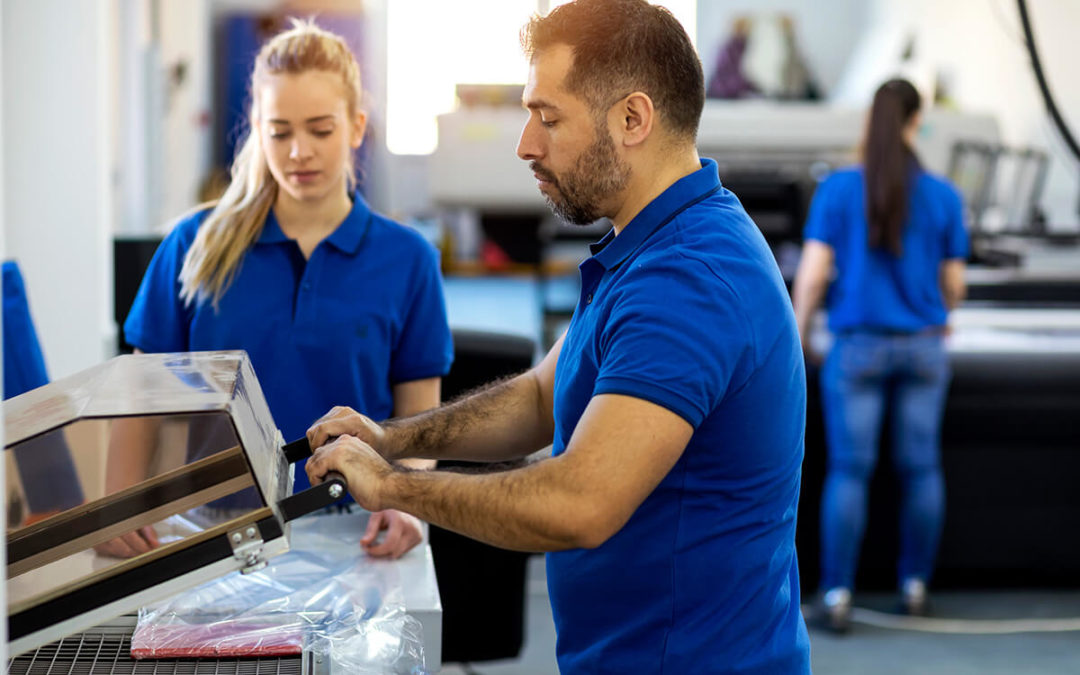 Two workers in blue shirts using a printing press in a branded apparel franchise workshop.