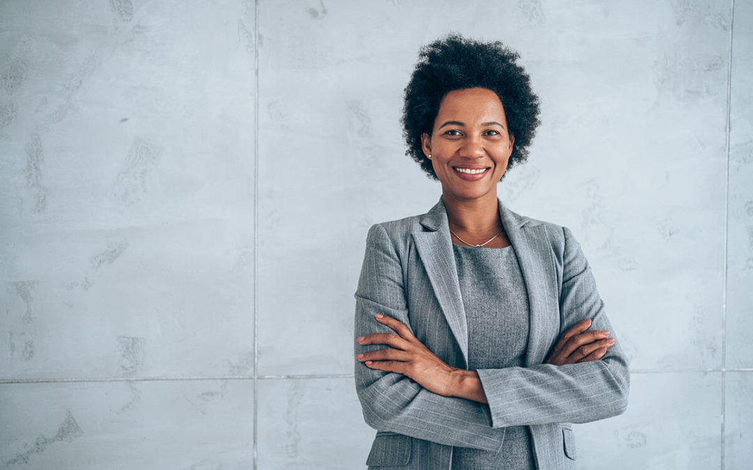 A confident black woman in a gray suit stands with arms crossed, smiling, representing a branded apparel franchise.