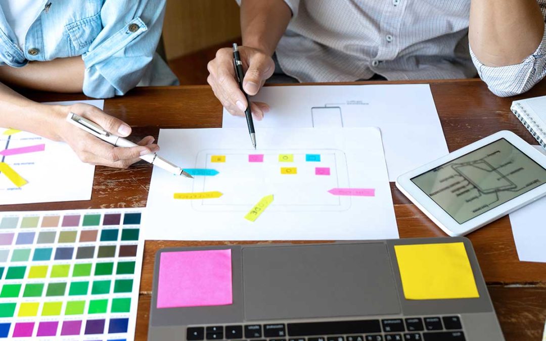 Dos personas colaborando en una mesa de madera, dibujando el diseño del sitio web de una franquicia de productos promocionales con marcadores de colores, notas adhesivas dispersas y herramientas de diseño.