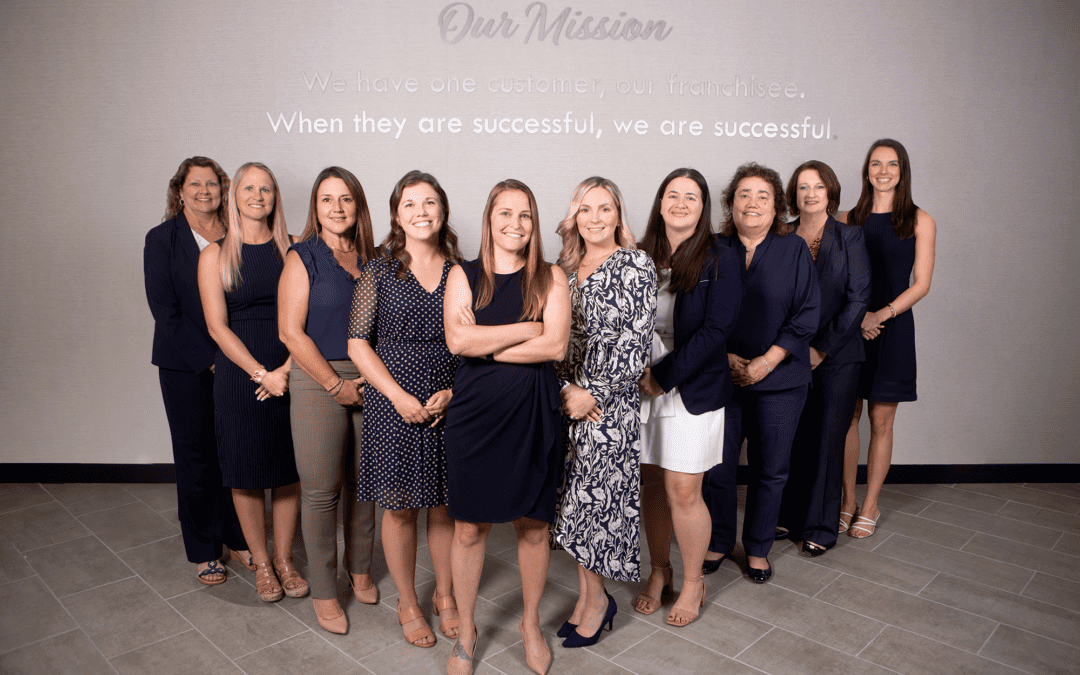 Diez mujeres profesionales paradas en fila en una oficina, sonriendo, cada una vistiendo uniformes de franquicias de ropa de marca.