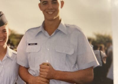 Un joven cadete militar con uniforme y gorra azul claro, sonriendo a la cámara, sosteniendo un bastón amarillo de franquicia de productos promocionales.