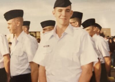 A young male military recruit in uniform smiling at the camera, surrounded by other recruits in a formation, part of a branded apparel franchise.