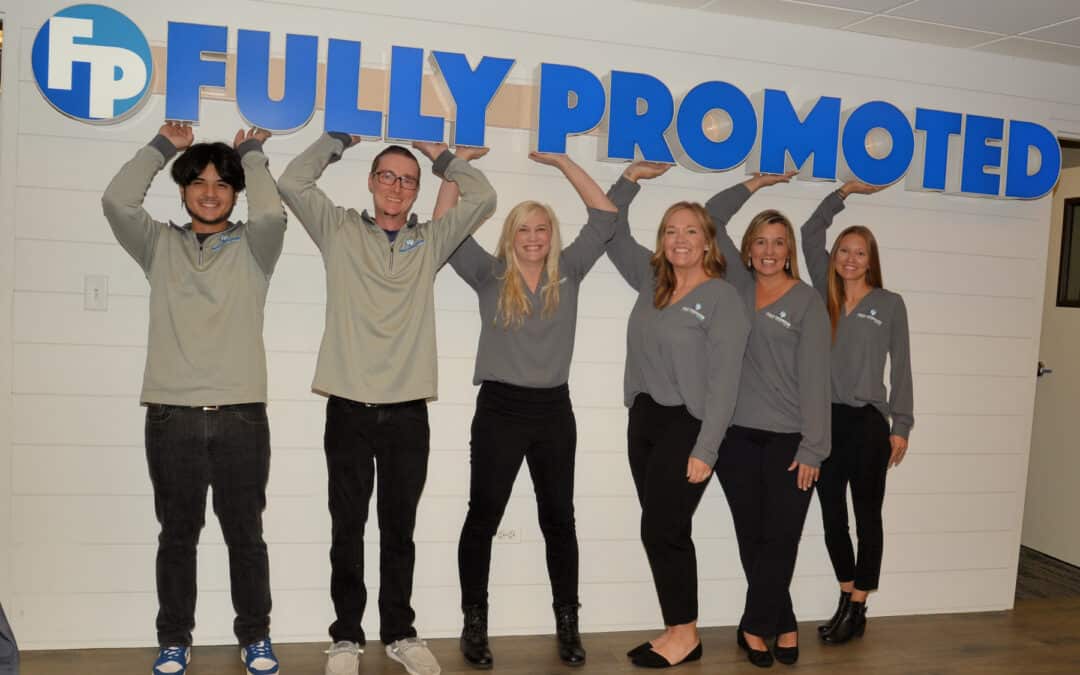 Five employees in gray shirts with the "fully promoted" branded apparel franchise logo pose with their arms raised in an office lobby.