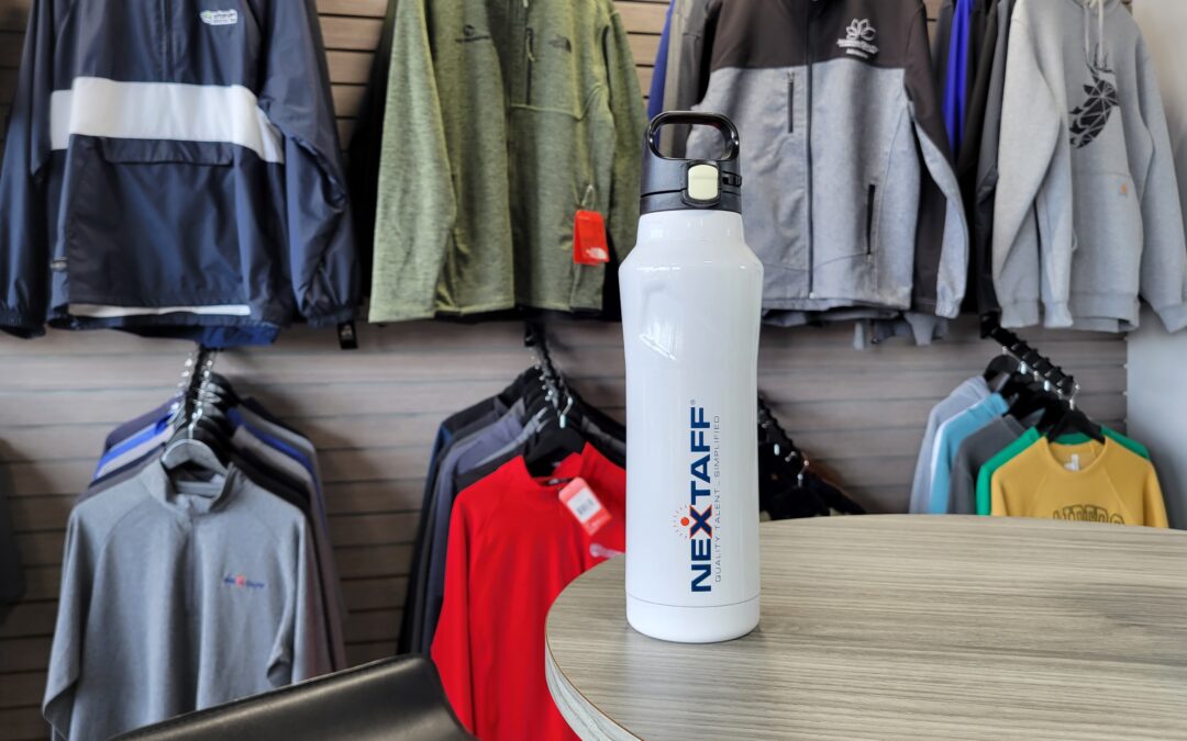 Una botella de agua blanca con el logo "nextaff" sobre una mesa, con varias chaquetas de colores colgadas en un perchero. Palabras clave no utilizadas: franquicia de ropa de marca, franquicia de productos promocionales.