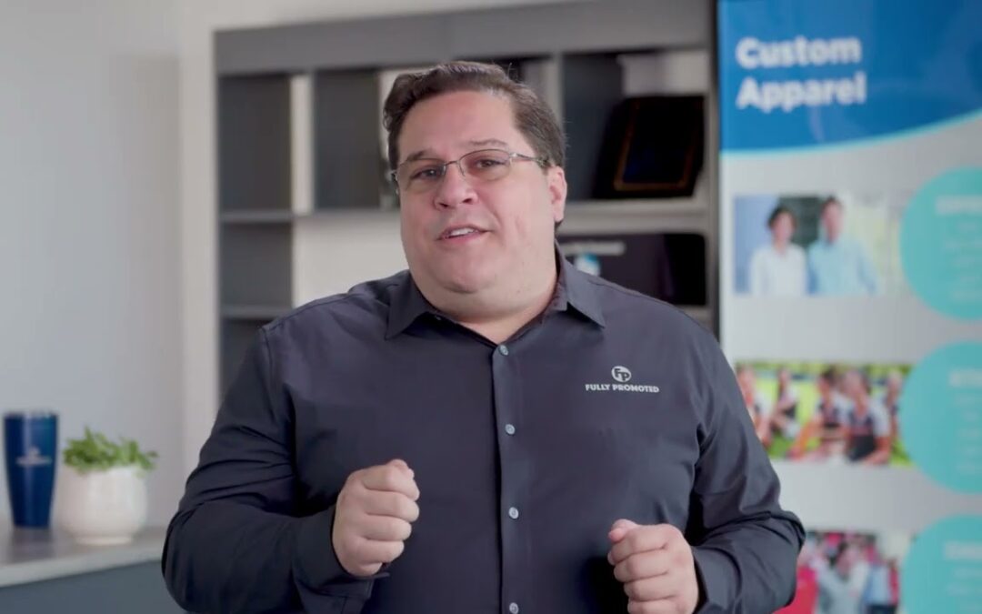 A man in a dark shirt speaking and gesturing, standing in an office environment with promotional product franchise posters.