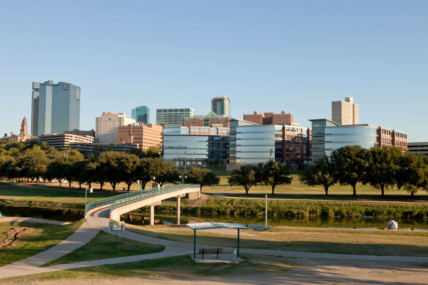 City skyline with modern and high-rise buildings beyond a park with a pathway, a bridge over a small body of water, trees, and an open shelter in the foreground.