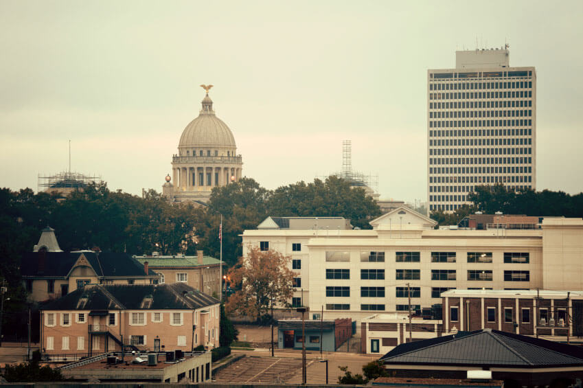 A cityscape featuring a domed capitol building, various structures, and a tall, modern office building. Trees and construction cranes are visible in the background under an overcast sky.