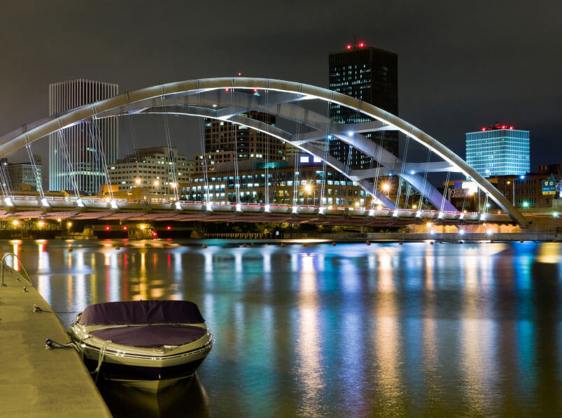 A modern arch bridge illuminated at night spans across a calm river with reflections, with a small boat docked in the foreground and city buildings in the background.