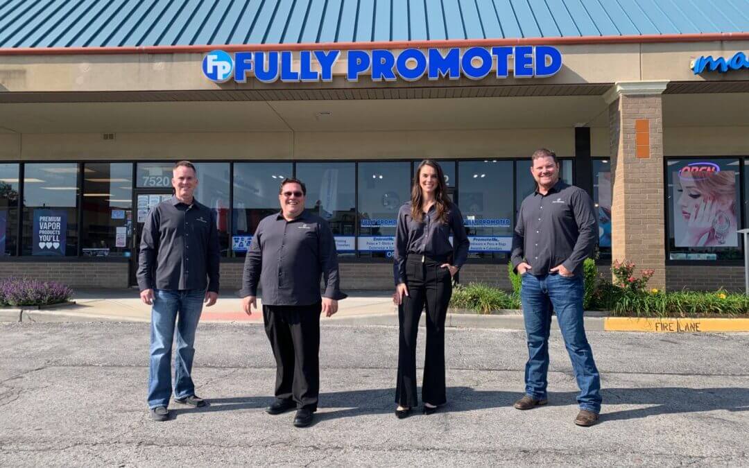 Four people stand in front of a store with a sign reading "Fully Promoted," wearing business casual attire. The store is part of a strip mall, with a sidewalk in front of them.