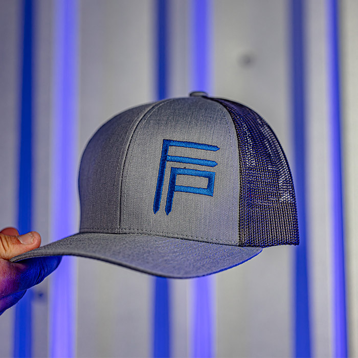 A hand holds a grey mesh-back baseball cap with a blue and teal "FP" logo against a metallic striped background.