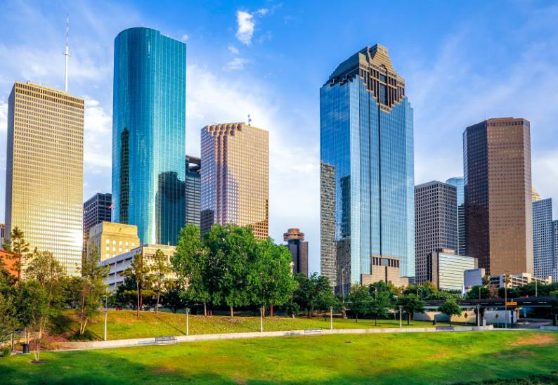 Skyline of modern skyscrapers in a city with a clear blue sky and green park in the foreground.