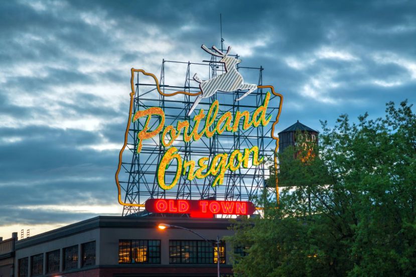 Neon sign reading "Portland Oregon" with a deer logo and "Old Town" at the bottom, set against a cloudy sky.